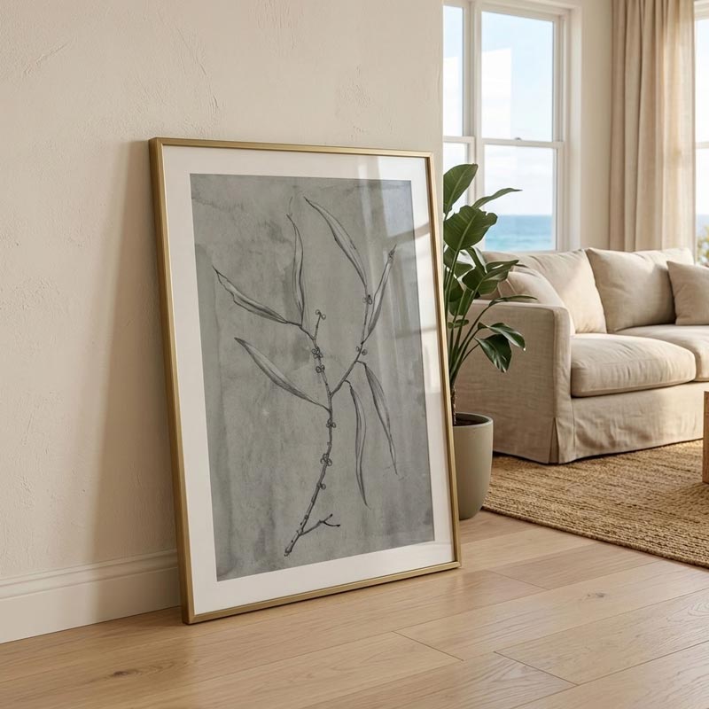 A framed botanical art print with a gold frame leans against a beige wall in a bright, modern living room with a sofa and potted plant.