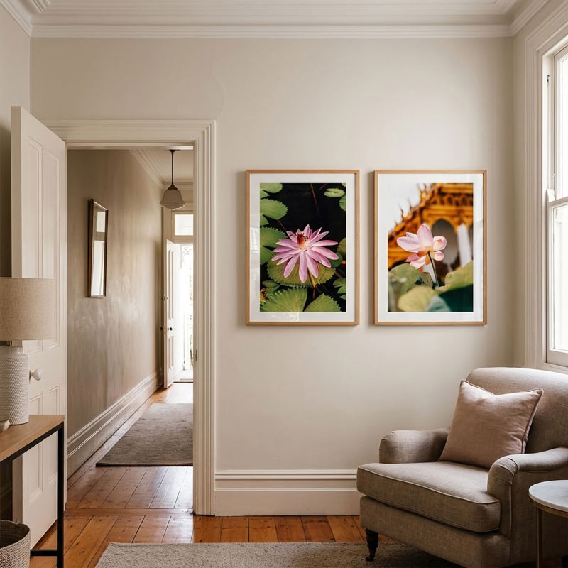 Two framed art prints depicting pink water lilies on a wall in a styled living room setting with a beige armchair, wooden floor, and hallway in the background.