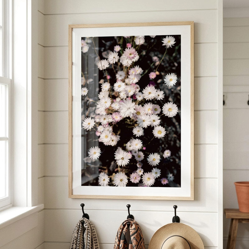 A framed floral art print featuring white and pale pink daisies on a dark background, hung on a white shiplap wall in a bright room with coat hooks and a straw hat below.