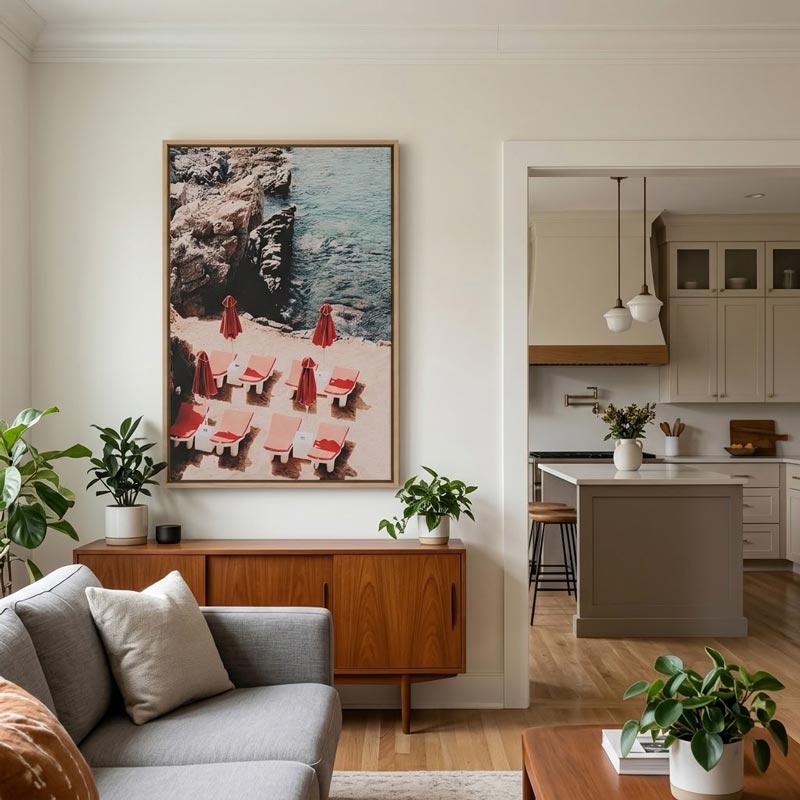 A framed vertical art print depicting a beach scene with red lounge chairs and umbrellas, displayed on a cream wall in a modern living room above a mid-century wooden sideboard.