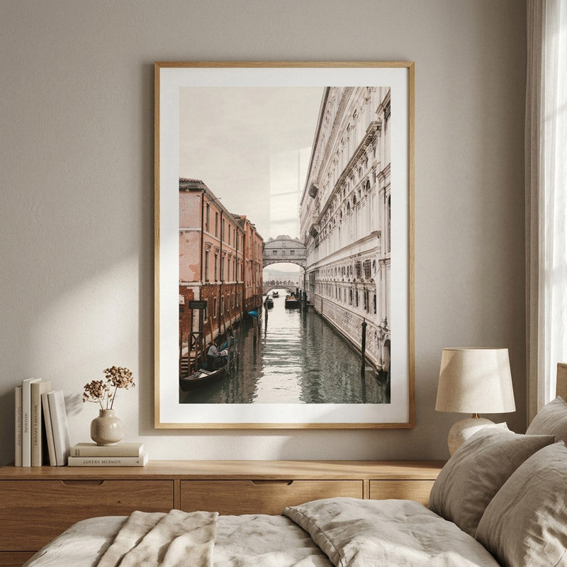 A framed vertical art print depicting a scenic canal with historic buildings, styled on a bedroom wall above a wooden nightstand with a lamp and books.