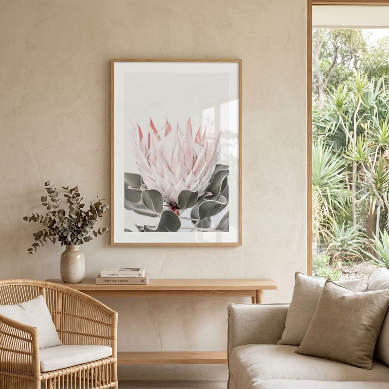 A framed botanical art print of a pink and gray protea flower hangs on a textured beige wall in a modern, sunlit living room with a wicker chair, sofa, and console table.