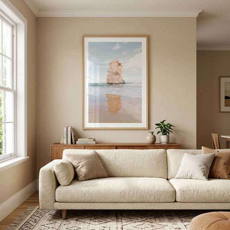 A framed vertical beach art print featuring a sea stack and reflection, displayed on a beige wall above a cream sofa in a styled living room.
