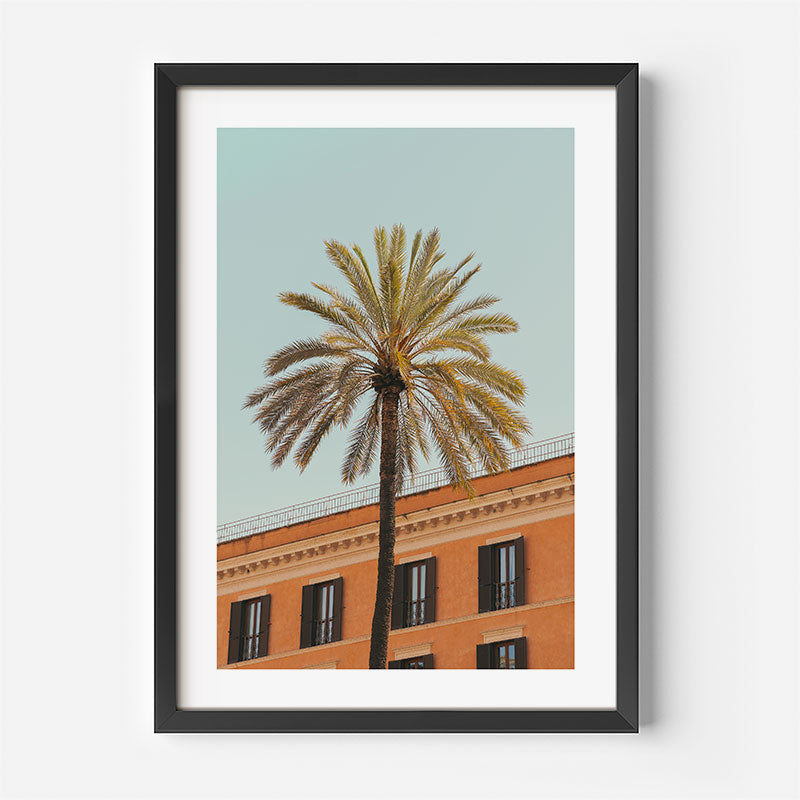 Framed wall art featuring a tall palm tree in front of a terracotta-colored building under a pale blue sky, shown in a black frame with a white mat against a light gray wall.