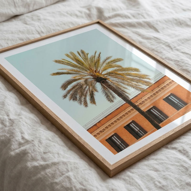 A framed art print featuring a palm tree against a light blue sky and an orange building, styled on a textured white linen surface.