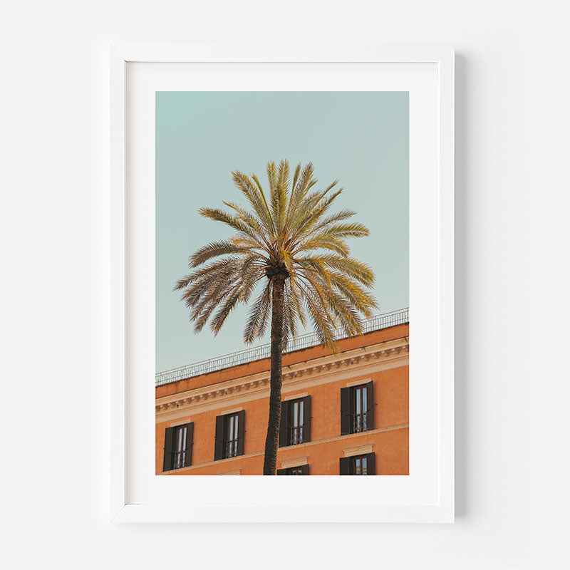 A framed art print featuring a tall palm tree against a pale blue sky and the top of a terracotta-colored building, displayed on a clean white wall.