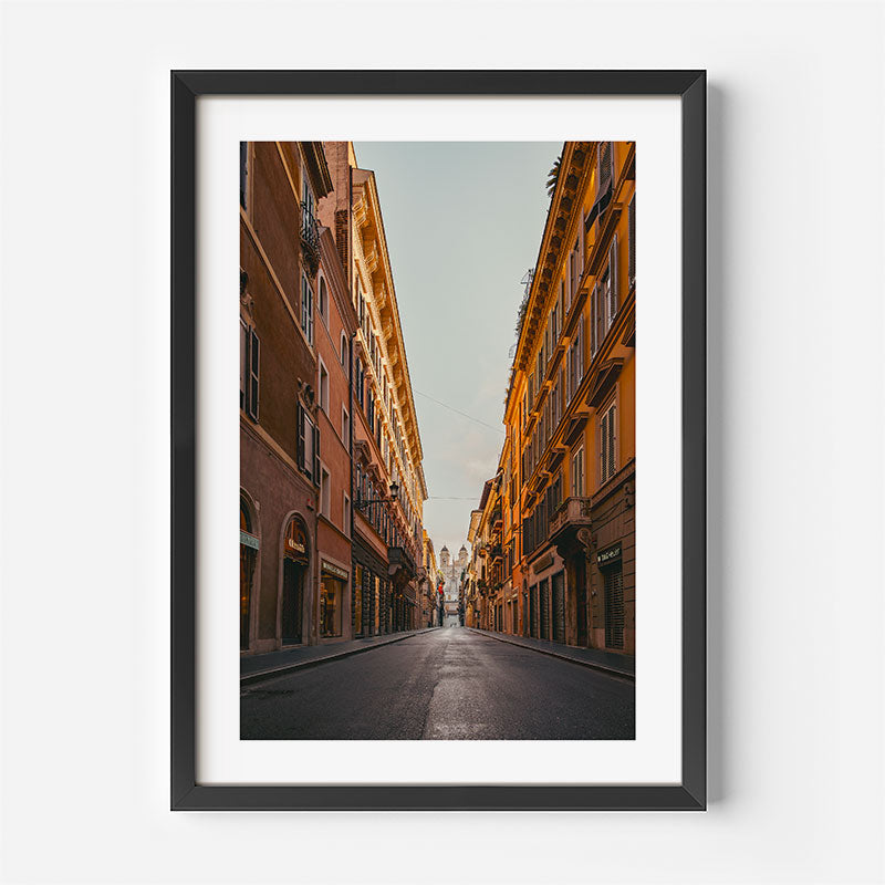 A framed vertical art print depicting a narrow city street lined with warm-toned historic buildings under a pale sky, shown in a black frame with a white mat against a light gray wall.