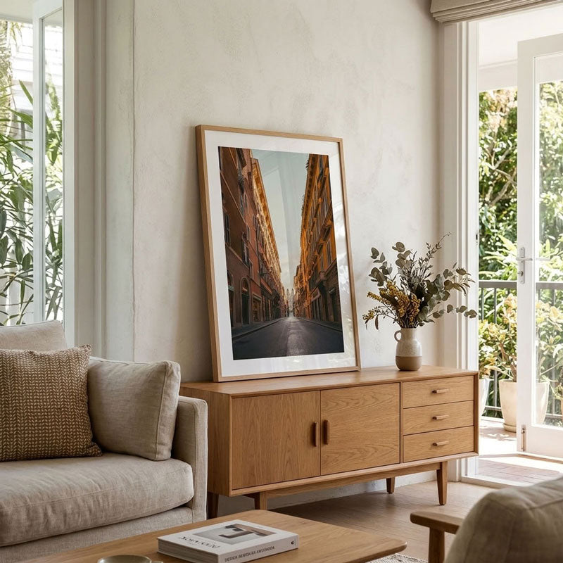 A large framed art print depicting a narrow city street lined with warm-toned buildings, styled in a light oak frame and leaning against a cream wall on a wooden sideboard in a modern living room.