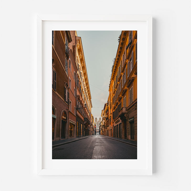 A framed vertical art print depicting a narrow city street lined with warm-toned buildings under a pale sky, presented in a white frame against a light gray wall.