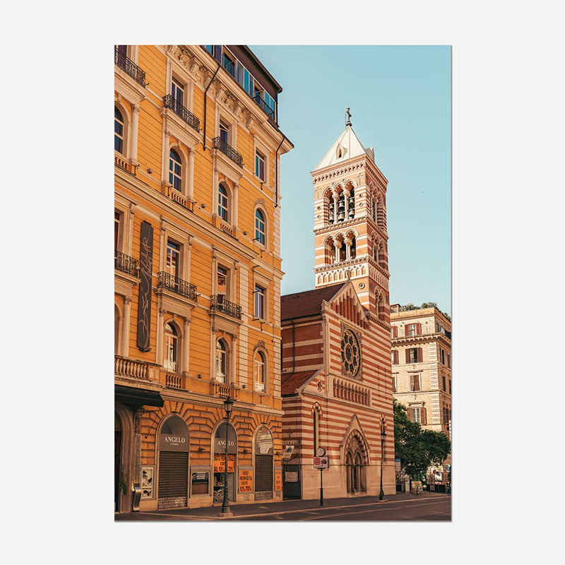 A vertical photographic print of a European street scene featuring a historic striped church tower and adjacent yellow-orange buildings under a clear blue sky, shown centered on a light gray background.