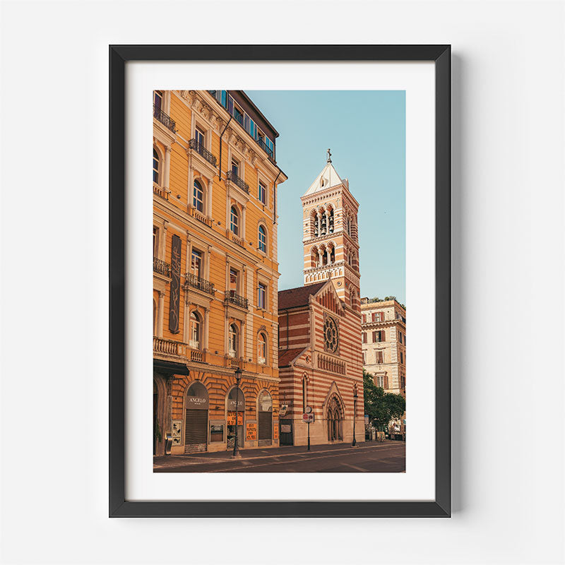 A framed art print featuring a photograph of a European street scene with a church tower and warm-toned buildings, displayed on a clean white background.