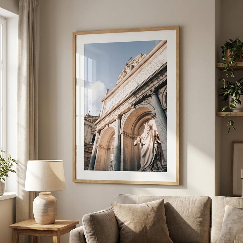 A large framed art print depicting a classical stone archway and statue, styled in a warm, modern living room setting above a beige sofa.