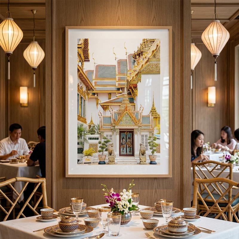 A large framed art print depicting a Thai temple courtyard with golden roofs and blue accents, hanging on a wooden wall in a well-lit restaurant with tables set for dining.
