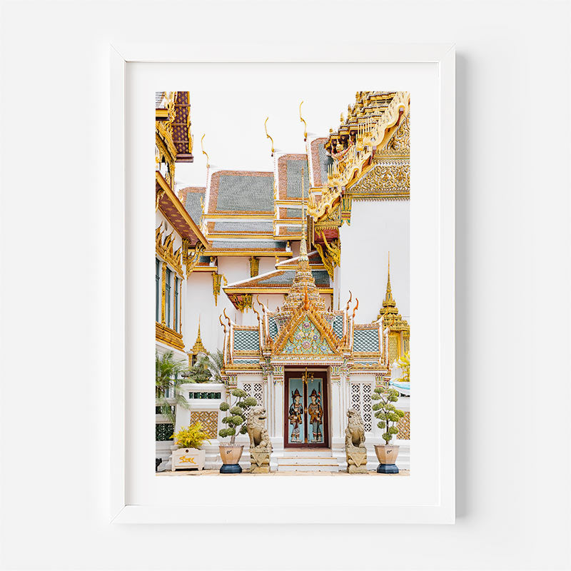 A framed art print featuring a photograph of a Thai temple courtyard with ornate golden roofs, white walls, and decorative statues, presented in a white frame against a light gray wall.