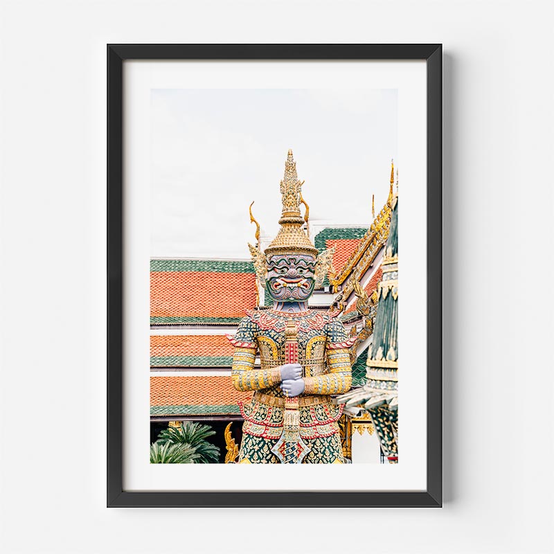 A framed art print of a Thai temple guard statue, displayed on a clean white wall. The print features a highly detailed, colorful statue with a blue face and ornate gold armor, set against a temple background with orange-red tiled roofs.