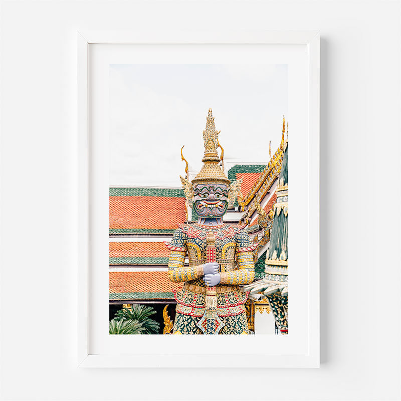 A framed art print featuring a colorful Thai temple guard statue with ornate gold and red roofs in the background, presented in a white frame against a light gray wall.