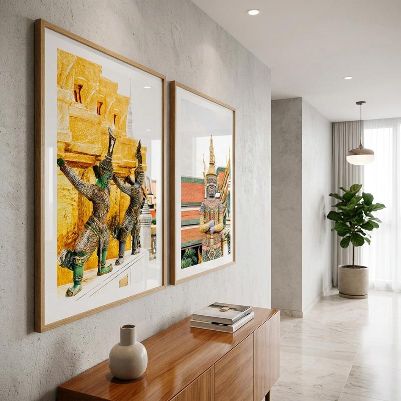 Two large framed art prints featuring golden Thai temple architecture and figures, displayed on a textured gray wall above a wooden console table in a modern, bright hallway.