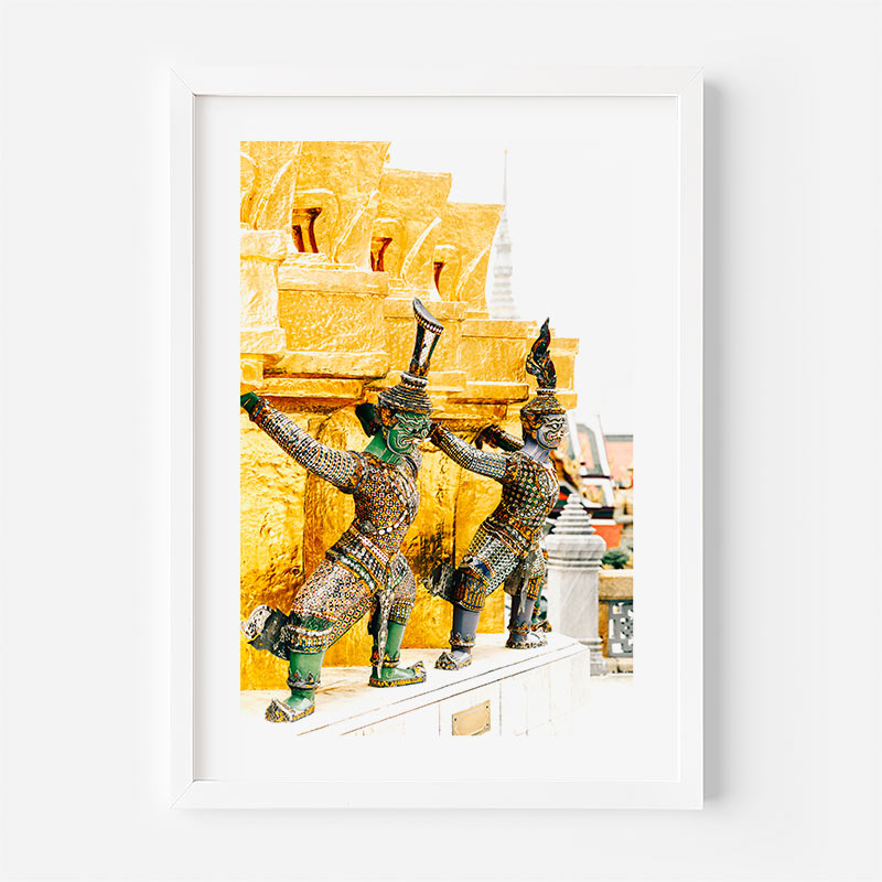 A framed art print featuring two Thai temple guards in green and gold mosaic armor against a golden background, presented in a white frame on a light gray wall.