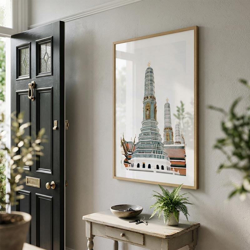 A framed art print depicting a Thai temple tower, styled on a light gray wall above a console table with a plant in a home entryway.