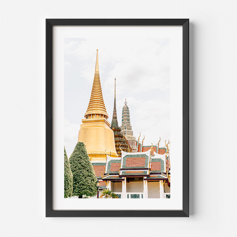 A framed art print of a Thai temple featuring a large golden chedi and traditional tiled roofs, presented in a black frame with a white mat against a light gray wall.
