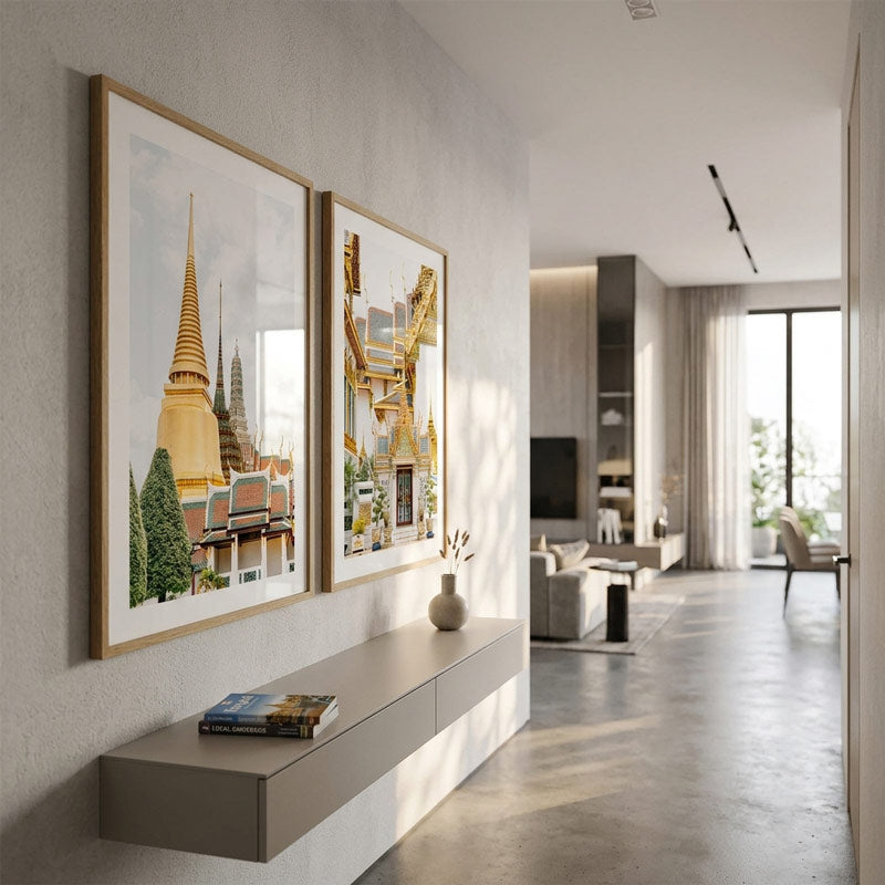 Two large framed art prints depicting golden temple spires and architectural details hang on a light gray wall in a modern, sunlit living room with a floating shelf and concrete floor.