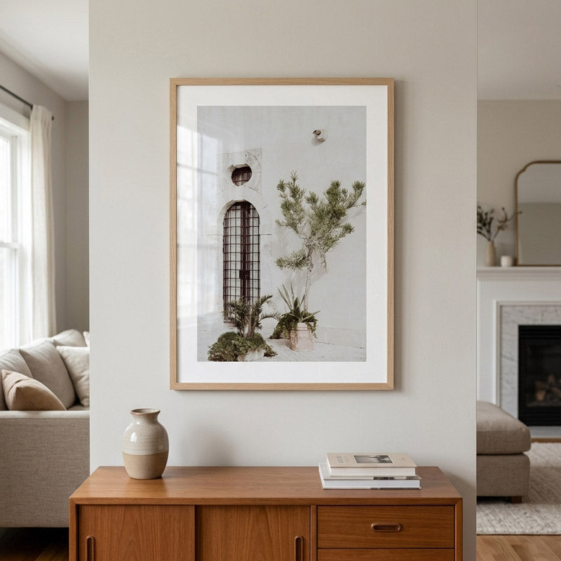 A framed botanical art print with a light wood frame and white mat, hanging on a beige wall above a wooden console table in a modern living room setting.