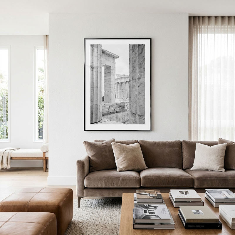 A black and white framed art print depicting ancient stone ruins, displayed on a white wall in a modern living room setting above a brown sofa.