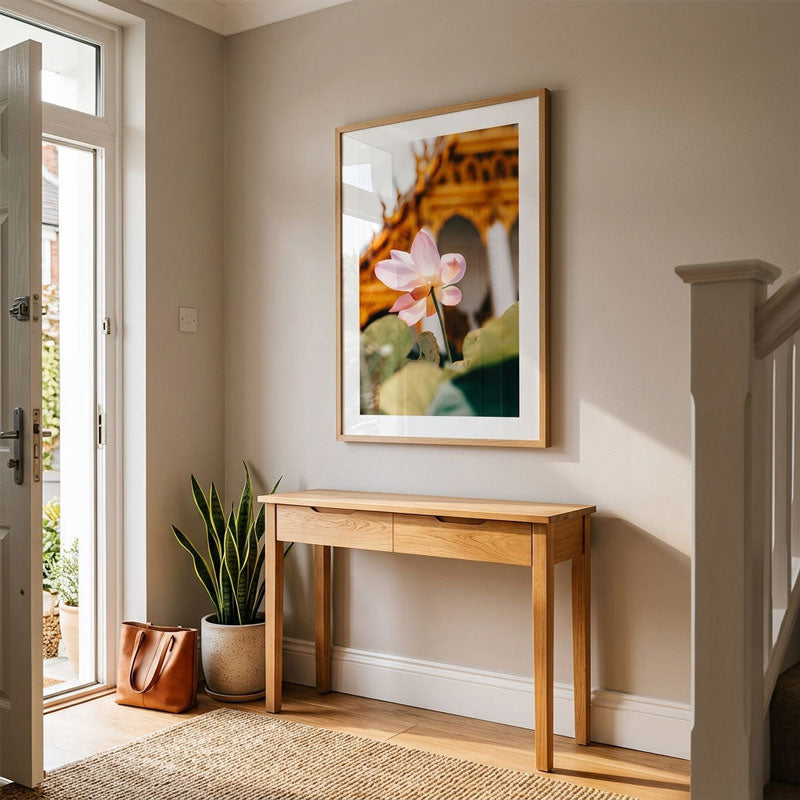 A framed art print featuring a pink lotus flower is displayed on a beige wall above a light wood console table in a bright, modern entryway.