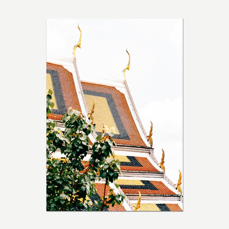 A vertical photographic print of a Thai temple roof with golden spires and patterned red, yellow, and black tiles, framed by green foliage in the foreground, shown against a light gray background.