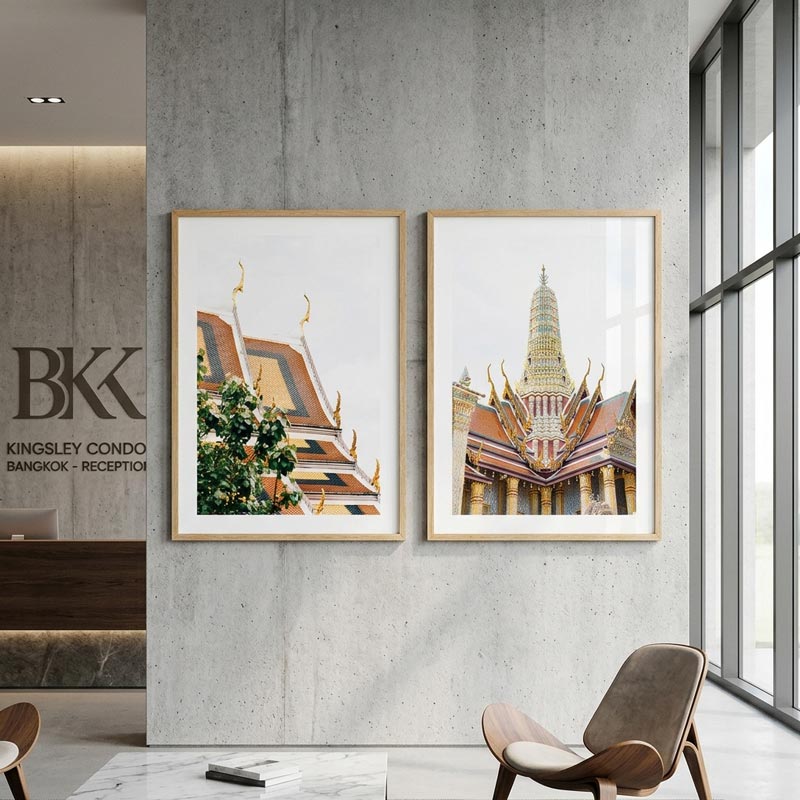 Two framed vertical art prints depicting Bangkok temple architecture hang on a textured gray concrete wall in a modern lobby setting. The left print shows a golden-roofed temple with green foliage, and the right print shows a tall spired temple tower.