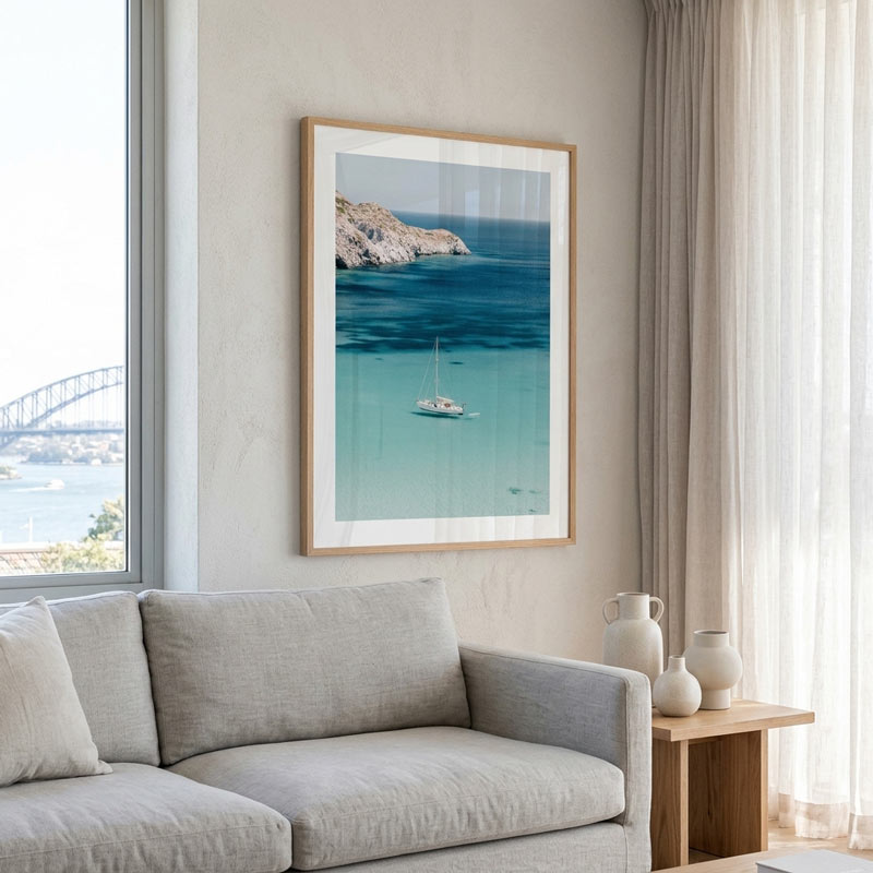 A framed vertical art print of a sailboat on turquoise water with a rocky island, displayed on a beige wall above a light gray sofa in a modern living room setting.