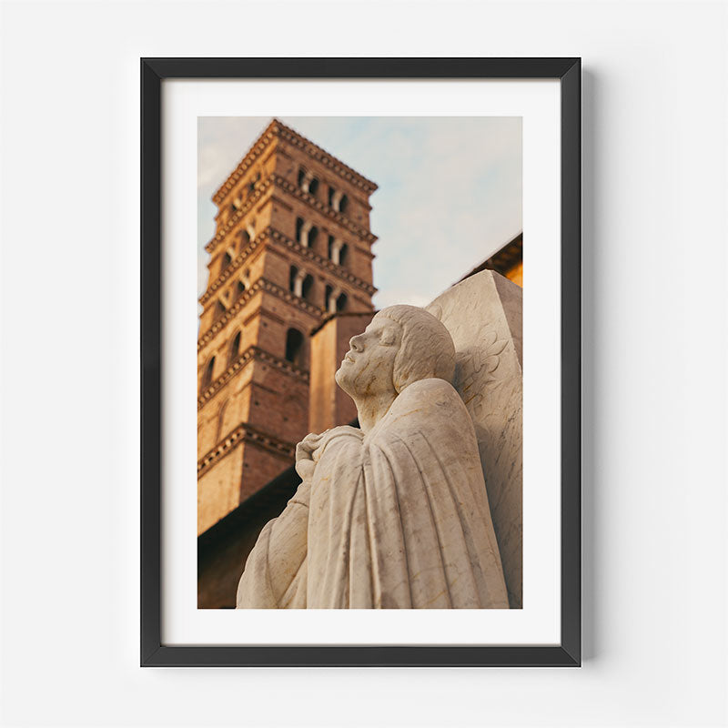Framed wall art featuring a photograph of a stone angel statue and a brick bell tower, shown in a black frame with a white mat against a light gray wall.