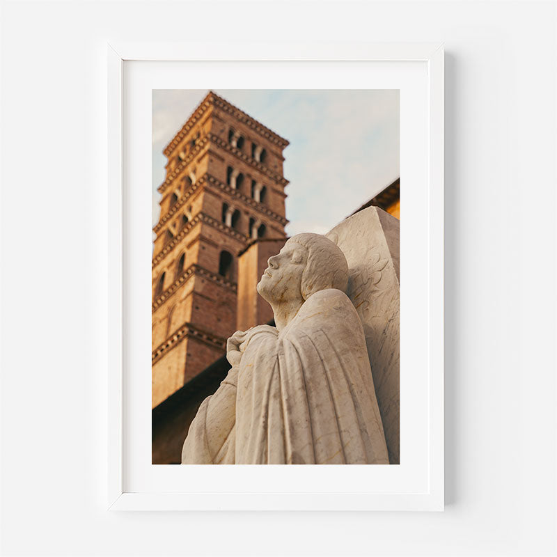 A framed art print featuring a low-angle photograph of a stone statue and a brick bell tower, presented in a white frame against a light gray wall.