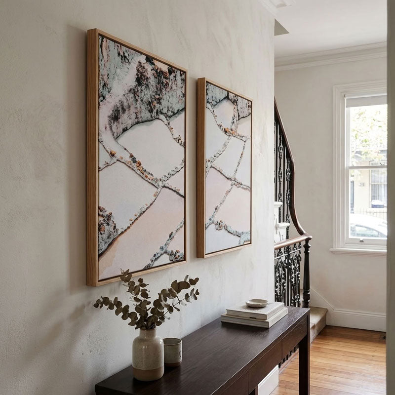 Two vertical framed abstract art prints with a marble-like pattern in black, gray, and soft pink tones, hanging on a textured off-white wall above a dark wood console table in a bright home interior.
