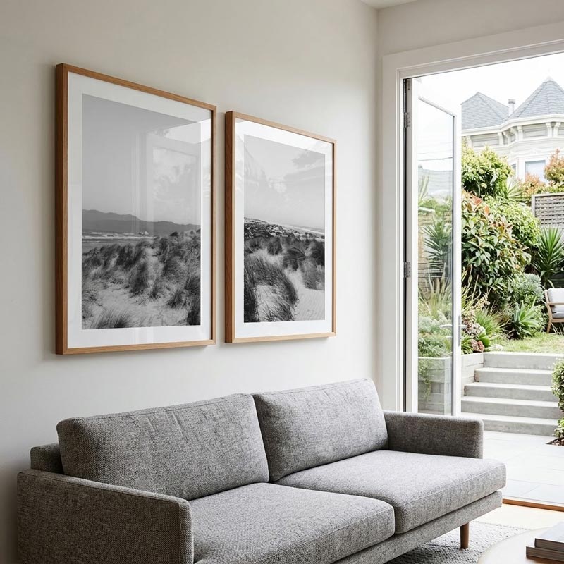 Two framed black and white beach dune photographs with light wood frames hang on a white wall above a gray fabric sofa, with an open glass door showing a garden and patio outside.