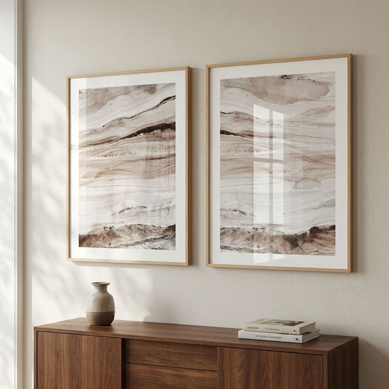Two framed abstract art prints with a marble-like pattern in beige, brown, and white tones, displayed side-by-side on a light beige wall above a wooden console table.