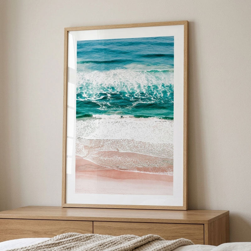 A framed vertical art print depicting turquoise ocean waves rolling onto a pale pink beach, displayed on a wooden dresser against a beige wall.