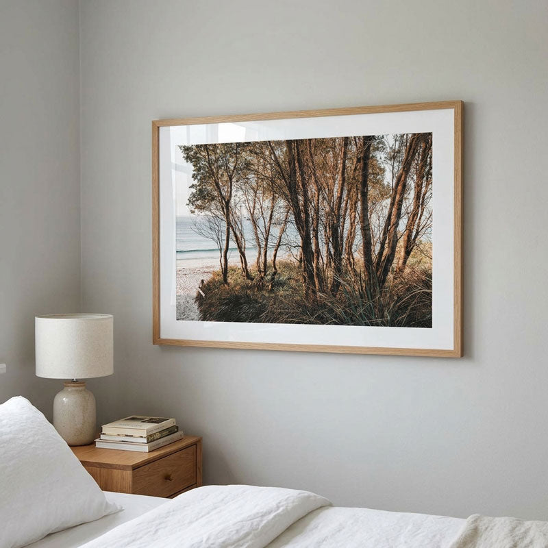A framed landscape photograph of a forest shoreline with trees, beach, and ocean, displayed on a light gray wall above a bed with white bedding and a wooden bedside table with a lamp.