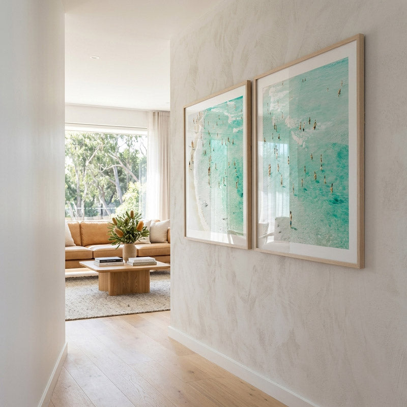 Two framed art prints depicting people at Bondi Beach hung on a wall in the hall way connecting to living room. The artwork captures the essence of a sunny beach day with a grainy texture and turquoise green and orange tones.