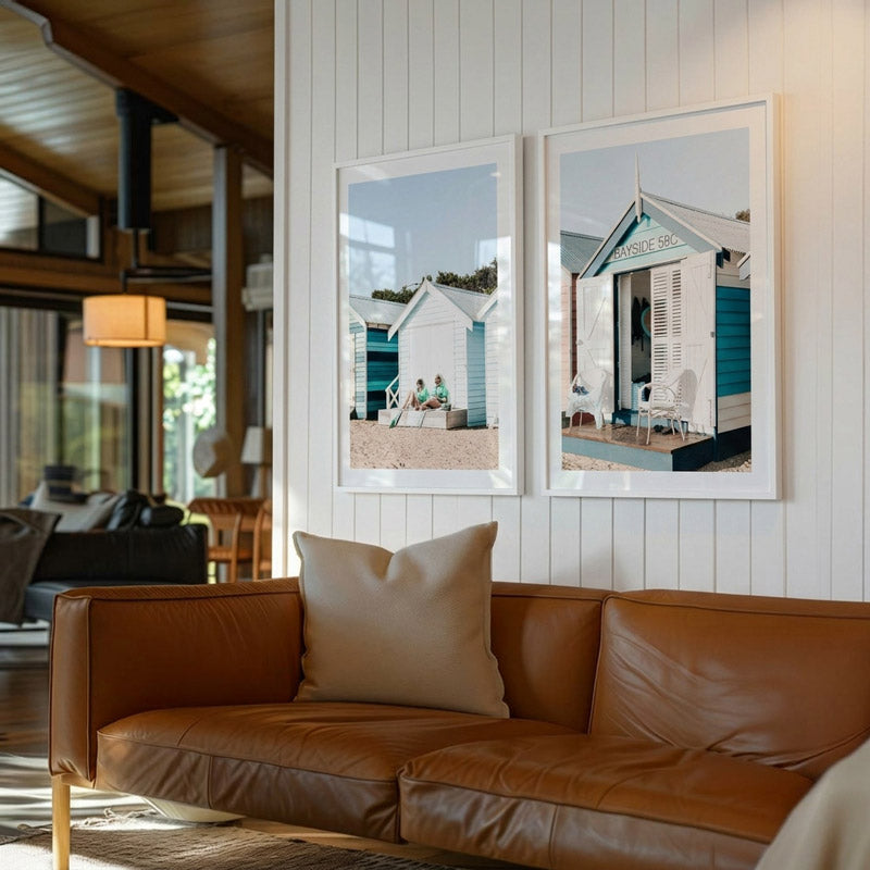 Two framed pictures of beach huts on a wall above a brown leather sofa.