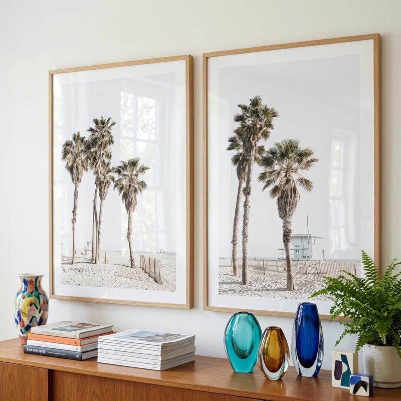 Two framed palm tree prints on a wall above a wooden shelf with decorative items.