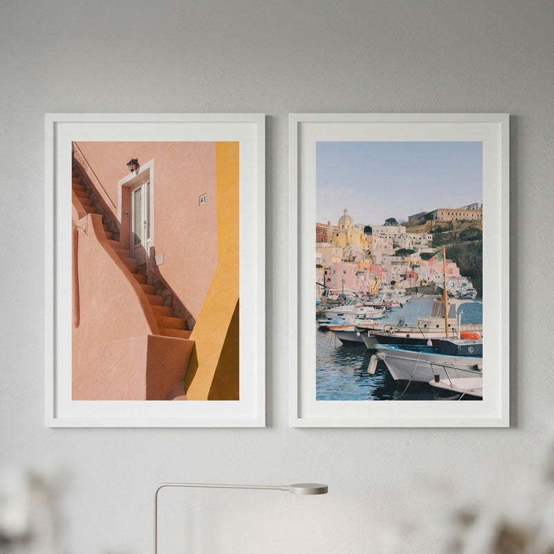 Two framed art prints hanging side-by-side on a light gray wall. The left print features a warm peach and yellow architectural scene with stairs, and the right print shows a colorful coastal harbor with boats and buildings.