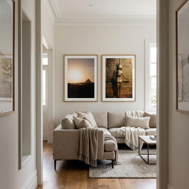 A modern living room with a beige sectional sofa, framed wall art, and hardwood floors, viewed through an open doorway. Two framed prints hang above the sofa, one showing a sunset over water and the other a bronze sculpture.