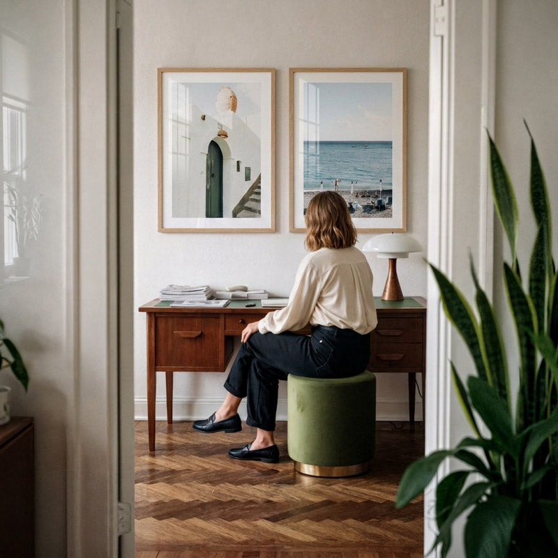Woman sitting on a green stool in a room with wooden floor and framed pictures on the wall.