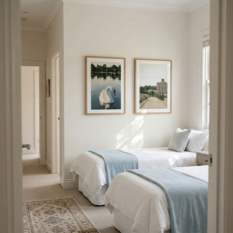 A bright, modern bedroom with two single beds dressed in white linens and light blue throws, featuring two framed landscape prints on the wall above. The room has off-white walls, a patterned area rug, and is viewed through a doorway.