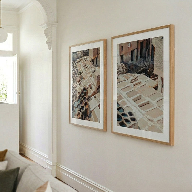 Two framed art prints with light wood frames and white mats hanging on a beige wall in a bright, modern room.