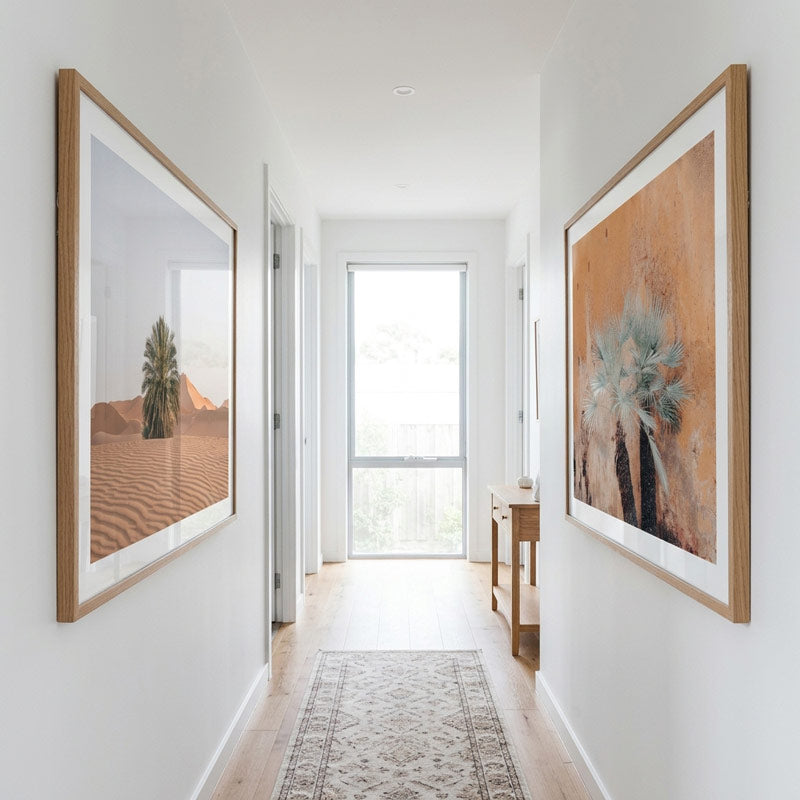 A long hallway with white walls, light wood flooring, a patterned runner rug, and two large framed wall art pieces. The left frame shows a desert landscape with a palm tree, and the right frame shows a close-up of palm trees with a warm orange-brown background.