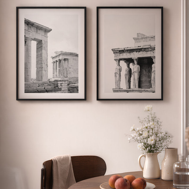 Two framed black and white architectural prints on a wall above a table with fruit and a vase.