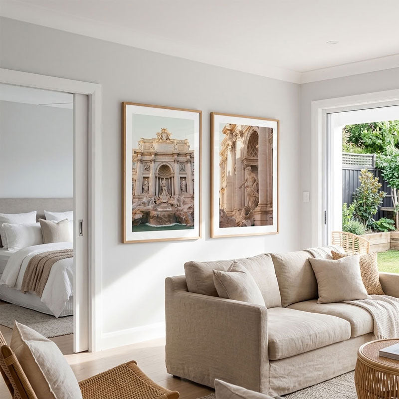 Two framed art prints depicting classical Roman architecture are displayed side-by-side on a light gray wall in a modern, sunlit living room with a beige sofa.