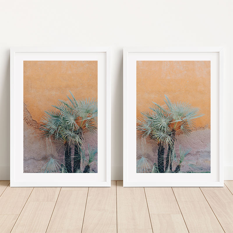 Two framed artworks of palm trees on a wooden floor with a white wall background